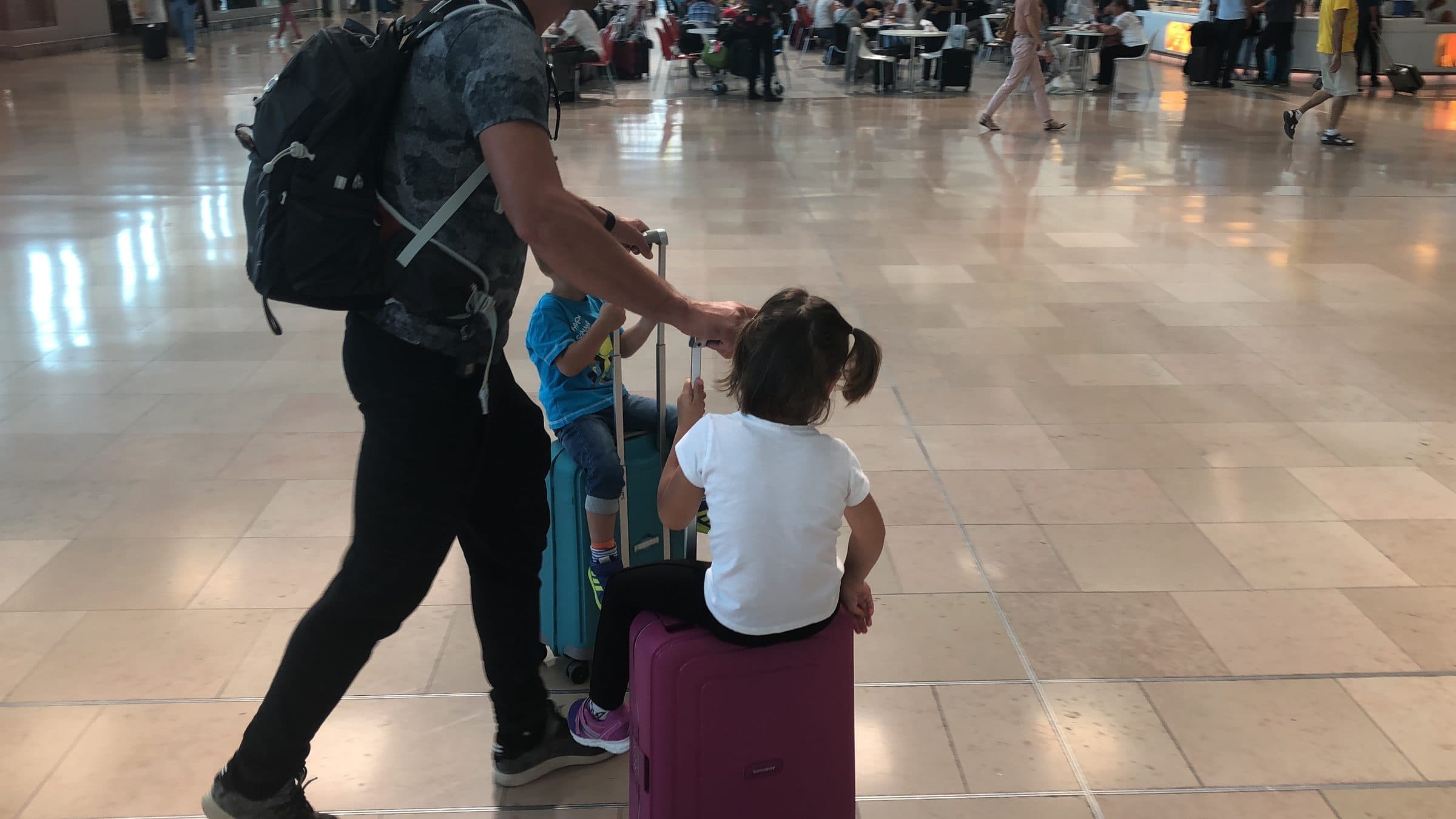 Two children sitting on suitcases at an airport, photographed from behind